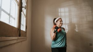 Senior woman in a brightly lit room exercises with dumbbells, promoting fitness at home.